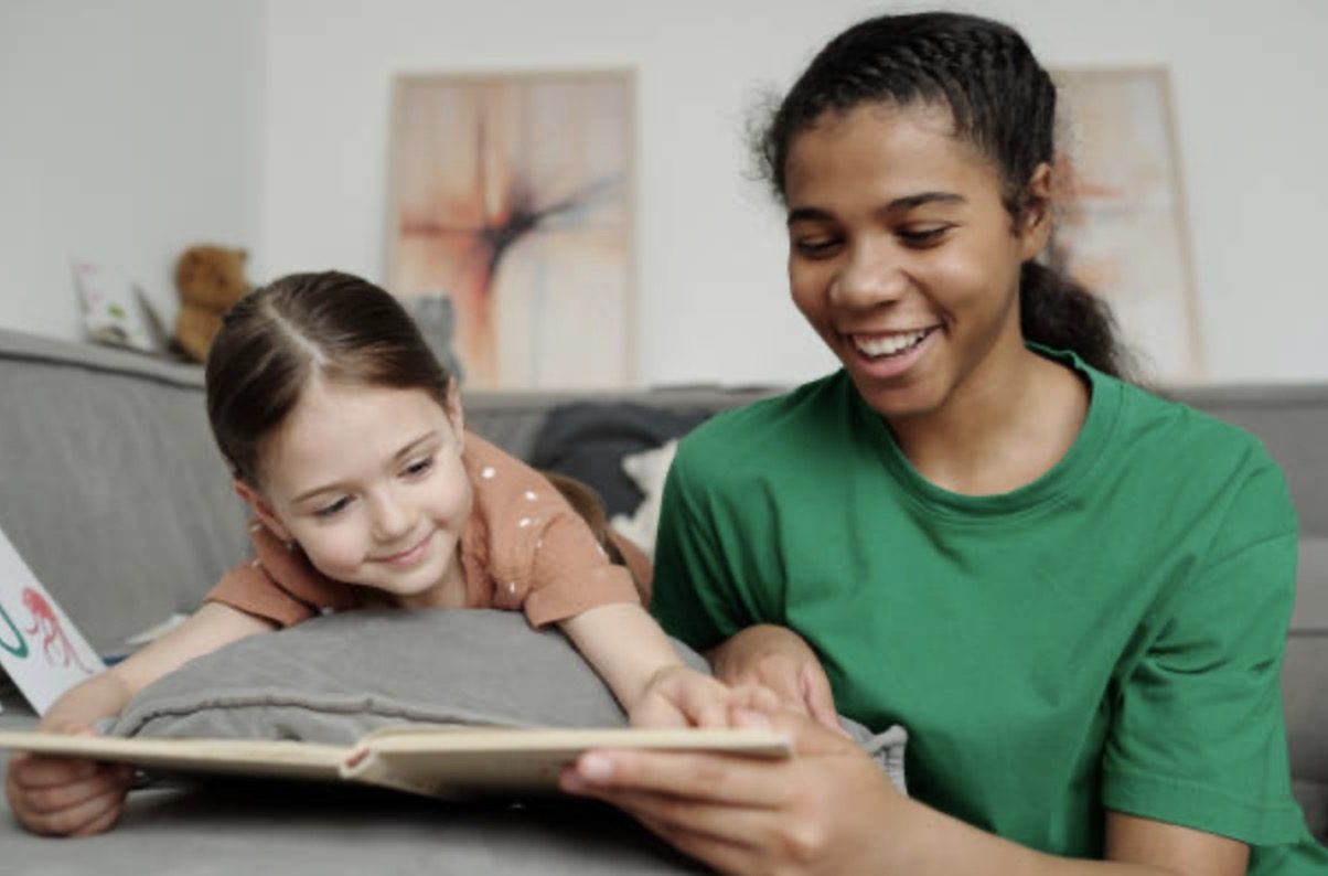 A smiling nanny reading a picture book with a young girl on the couch